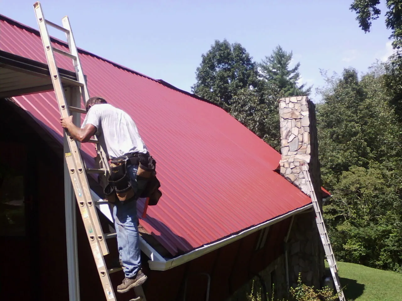 Roofer on a ladder beside a red metal roof and stone chimney — Soffit & Fascia Repair by Roofing Brothers in Rock River